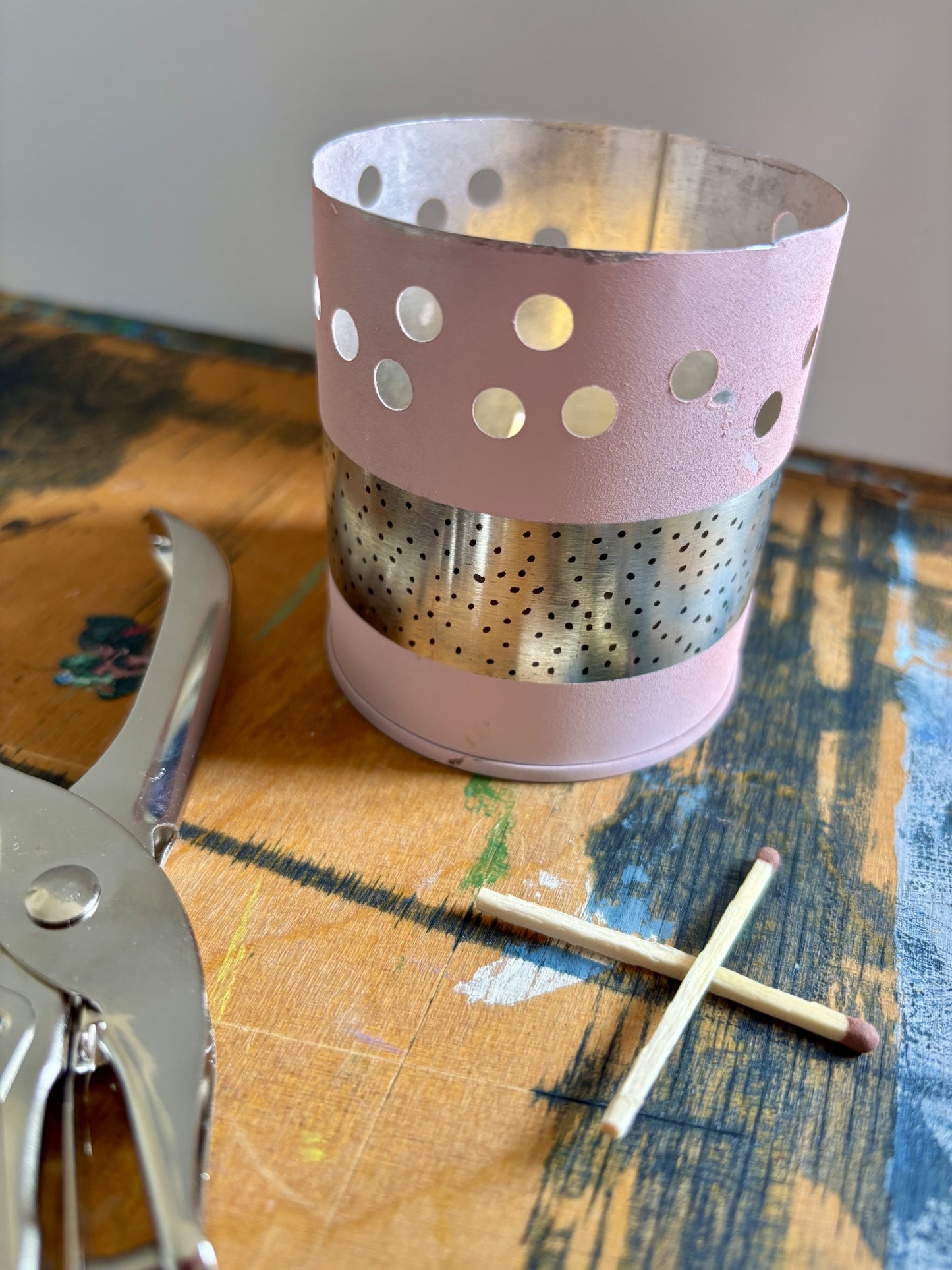 A pink metal candle holder with circular cutouts and a perforated silver band, shown on a painted wooden surface with a match and metal tool nearby.