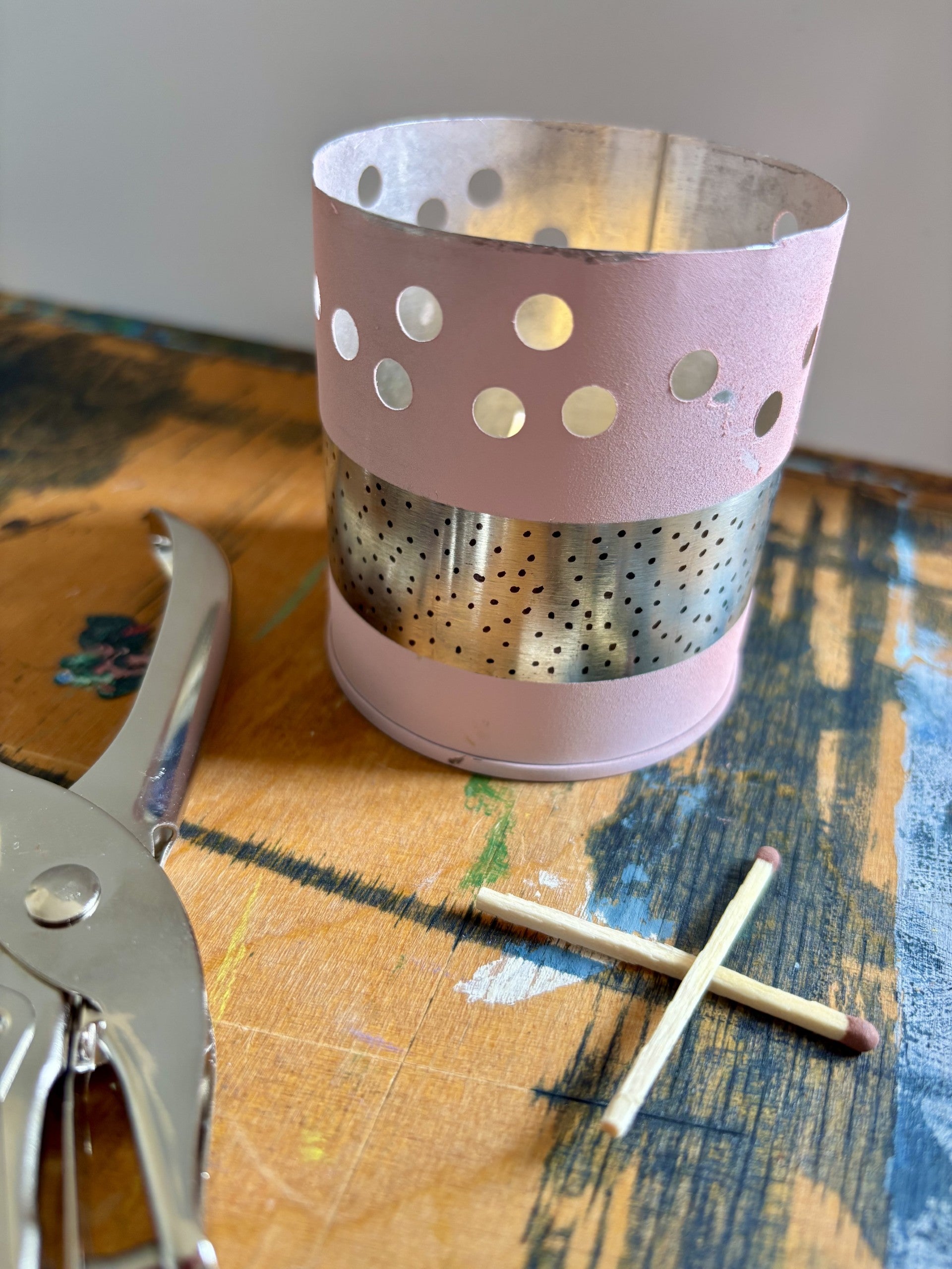 A pink metal candle holder with circular cutouts and a perforated silver band, shown on a painted wooden surface with a match and metal tool nearby.