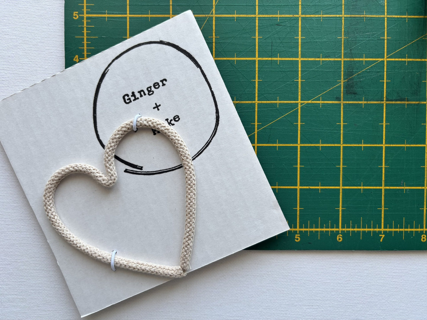 A handmade wire heart with beige cord, displayed on white card printed with 'Ginger + Make' next to a green cutting mat.