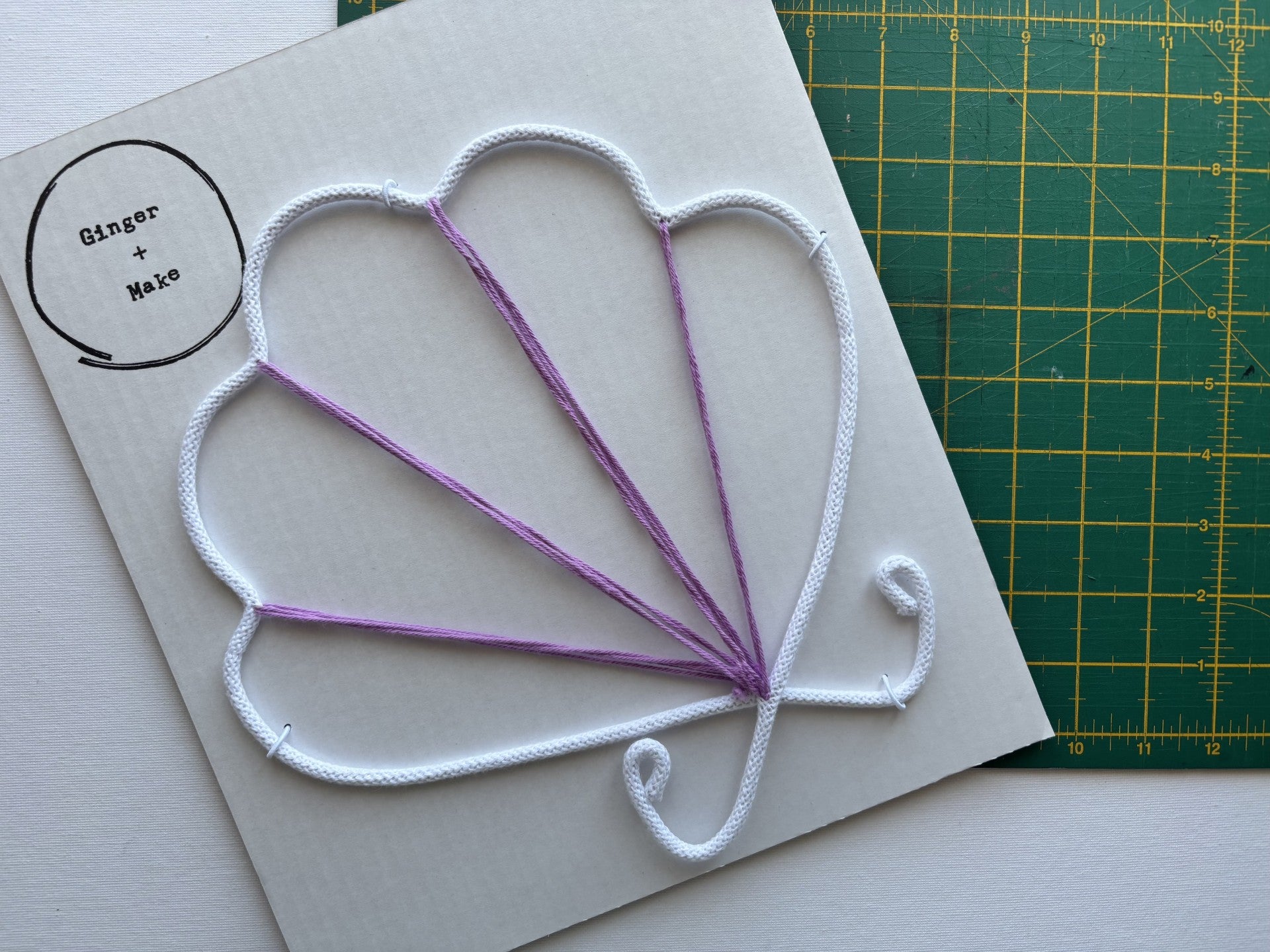 A handmade wire shell with white cotton cord outlining a scallop pattern and purple cotton forming the inner rays, placed on white card next to a green cutting mat. A circular logo in the top-left corner reads 'Ginger + Make'.