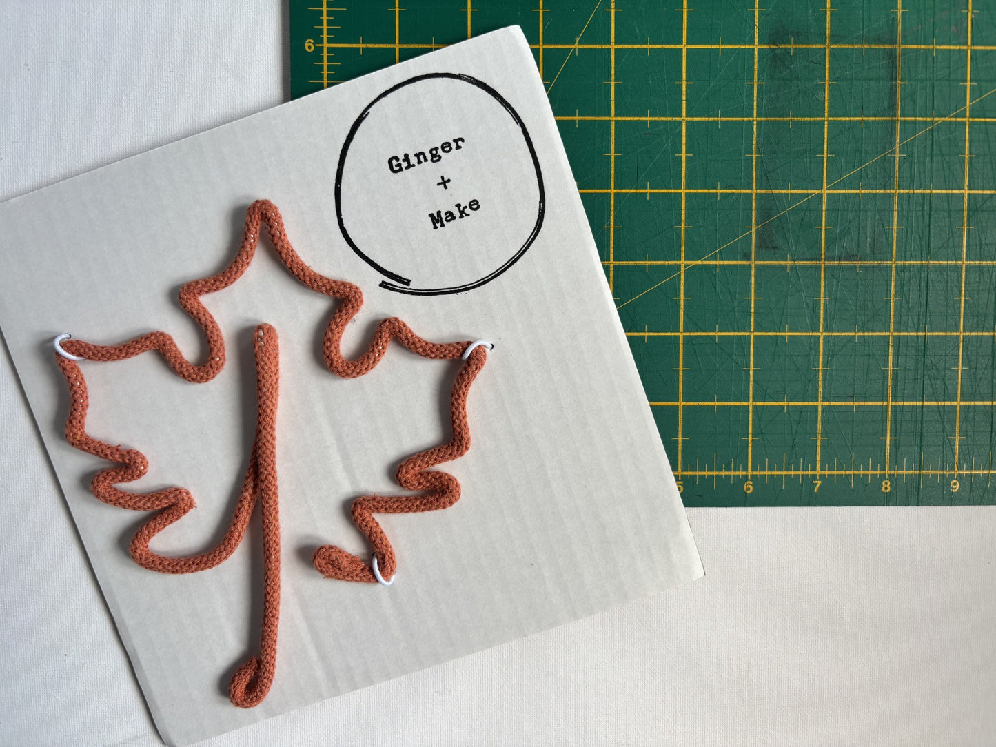 A white rectangular card with a wire maple leaf and a circular label reading 'Ginger + Make' sits on a green cutting mat with yellow grid lines. 