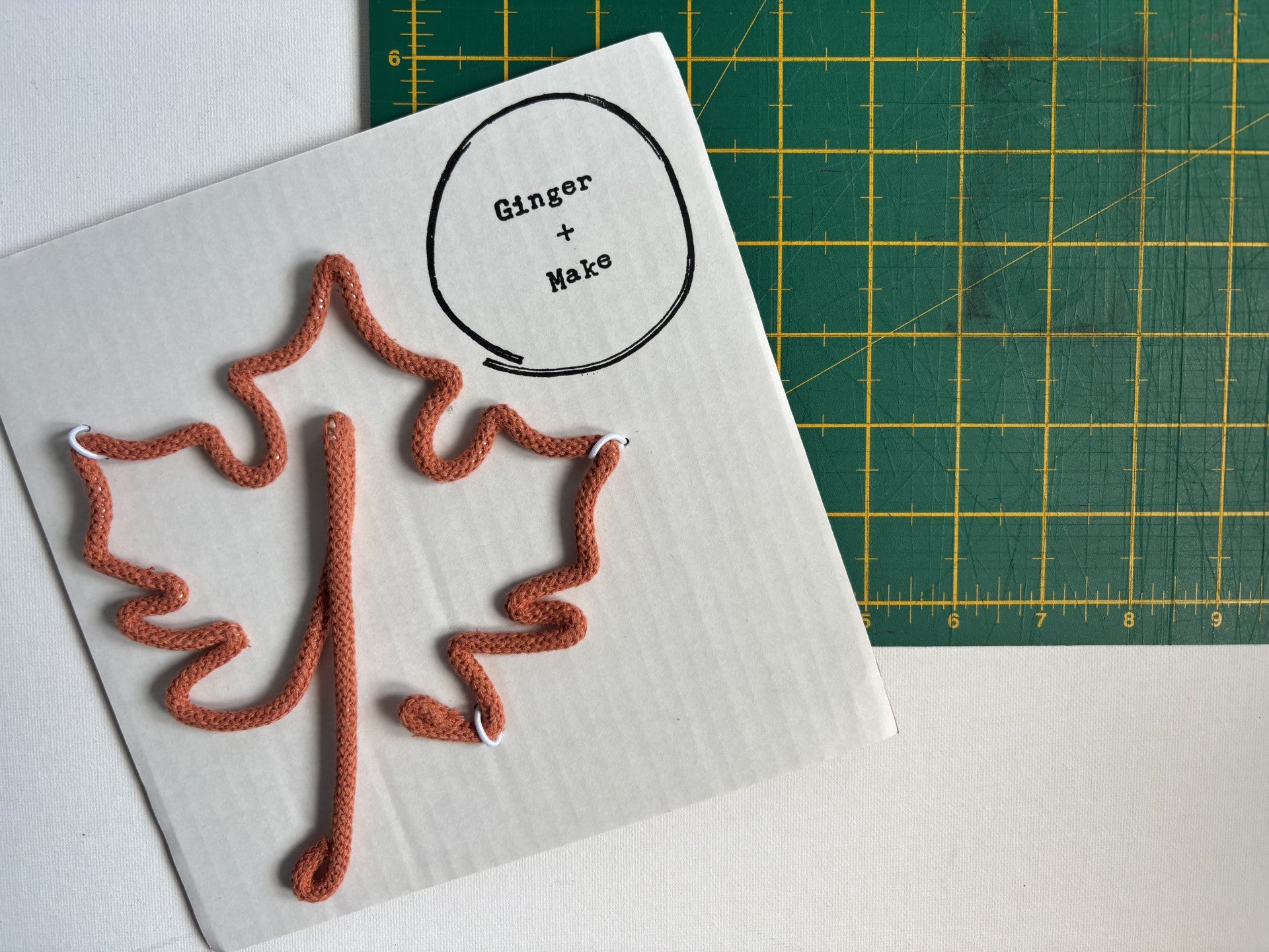 A white rectangular card with a wire maple leaf and a circular label reading 'Ginger + Make' sits on a green cutting mat with yellow grid lines. 
