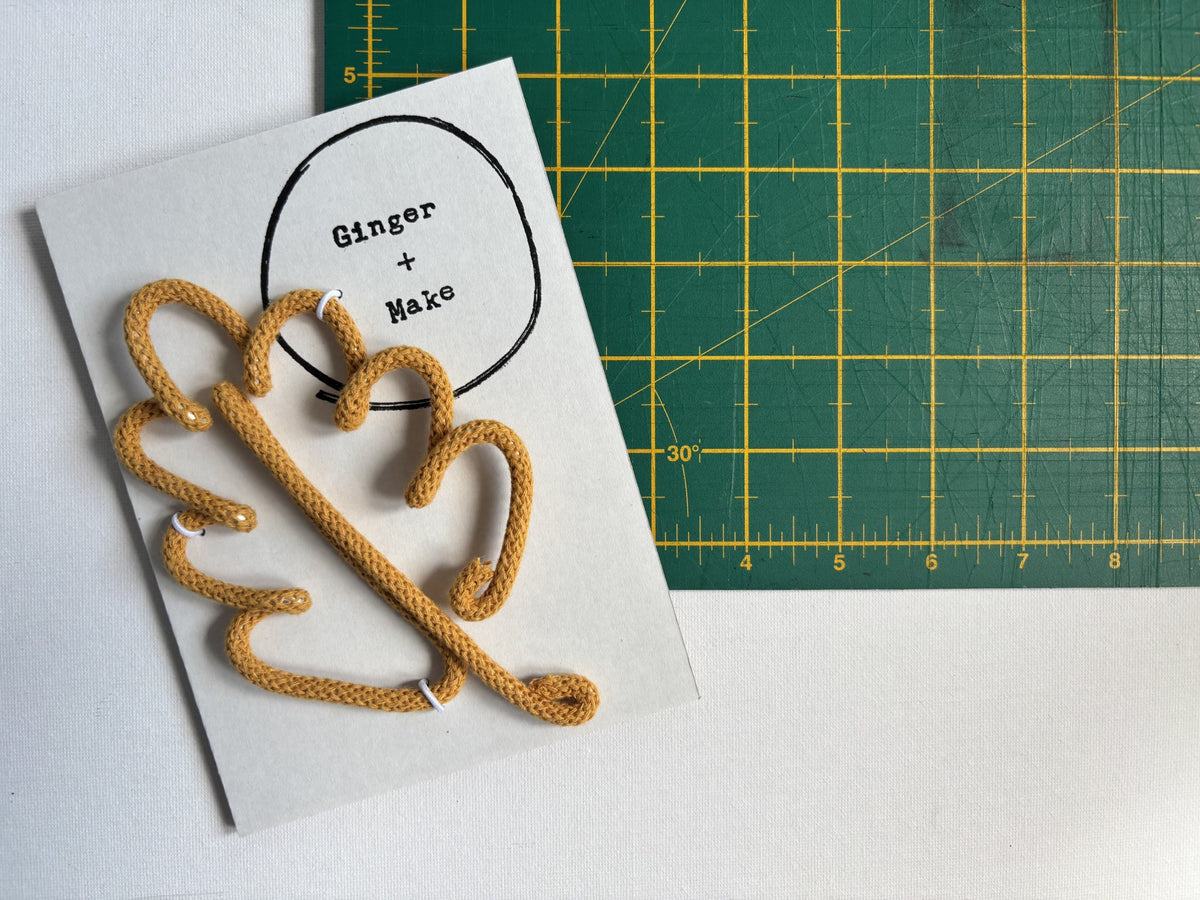 A mustard yellow wire and cord leaf on a white card with a printed logo reading 'Ginger + Make', placed on top of a green cutting mat with yellow grid lines.