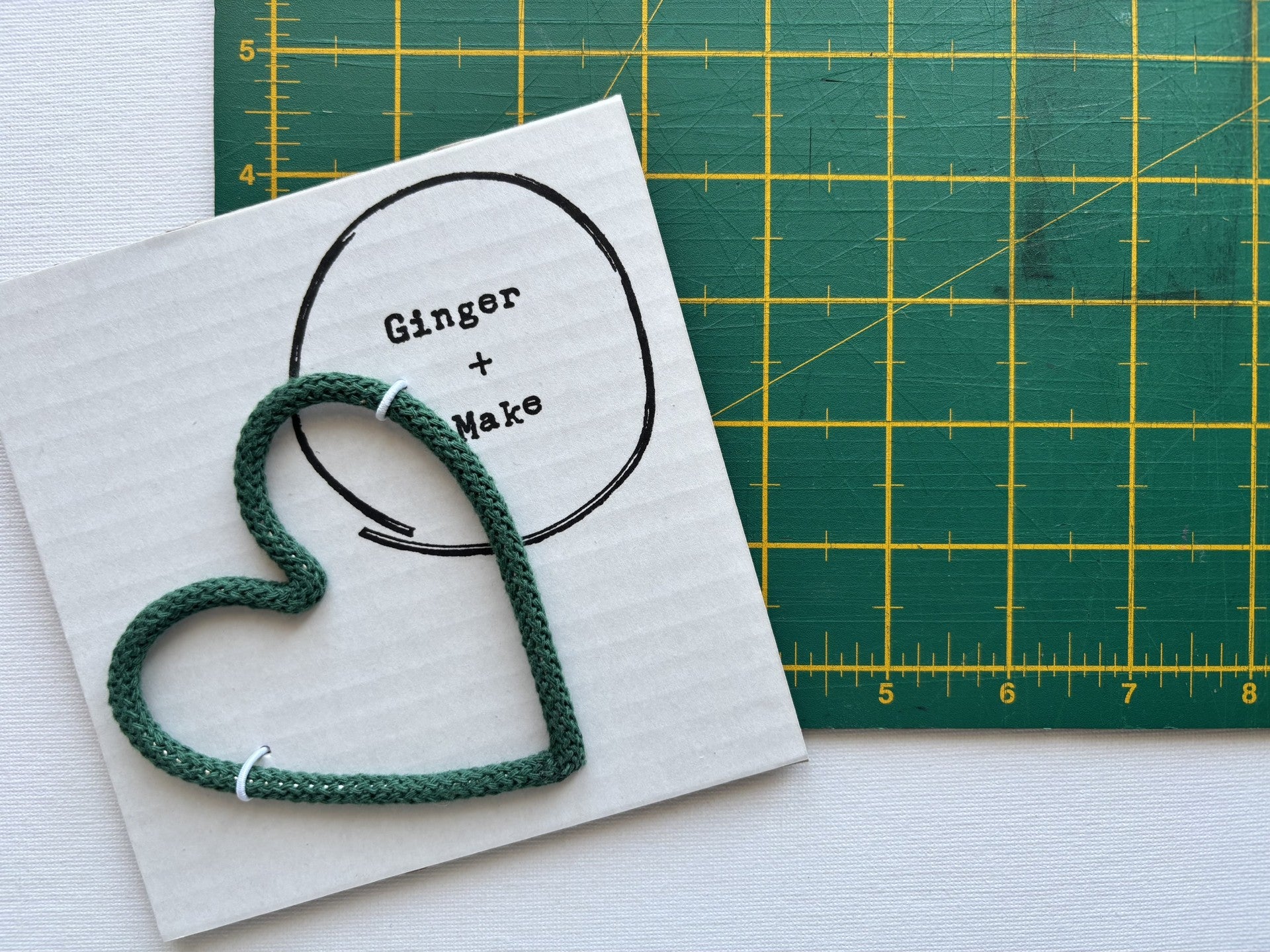 A handmade wire heart with green cord, displayed on white card printed with 'Ginger + Make' next to a green cutting mat.