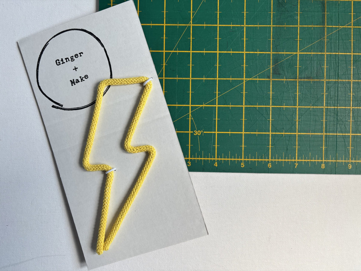 Wire and yellow cord shaped like a lightning bolt, attached to a white card printed with the words 'Ginger + Make', placed on a green cutting mat.