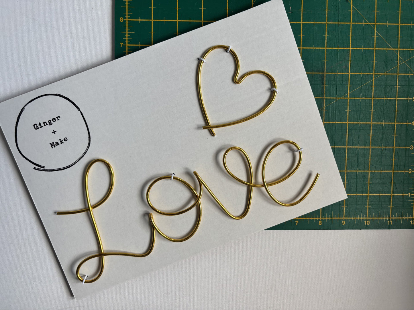 Gold-colored wire lettering spelling 'LOVE' with a heart on a white backing card, placed on a green cutting mat. A circular logo in the top-left corner reads 'Ginger + Make'.