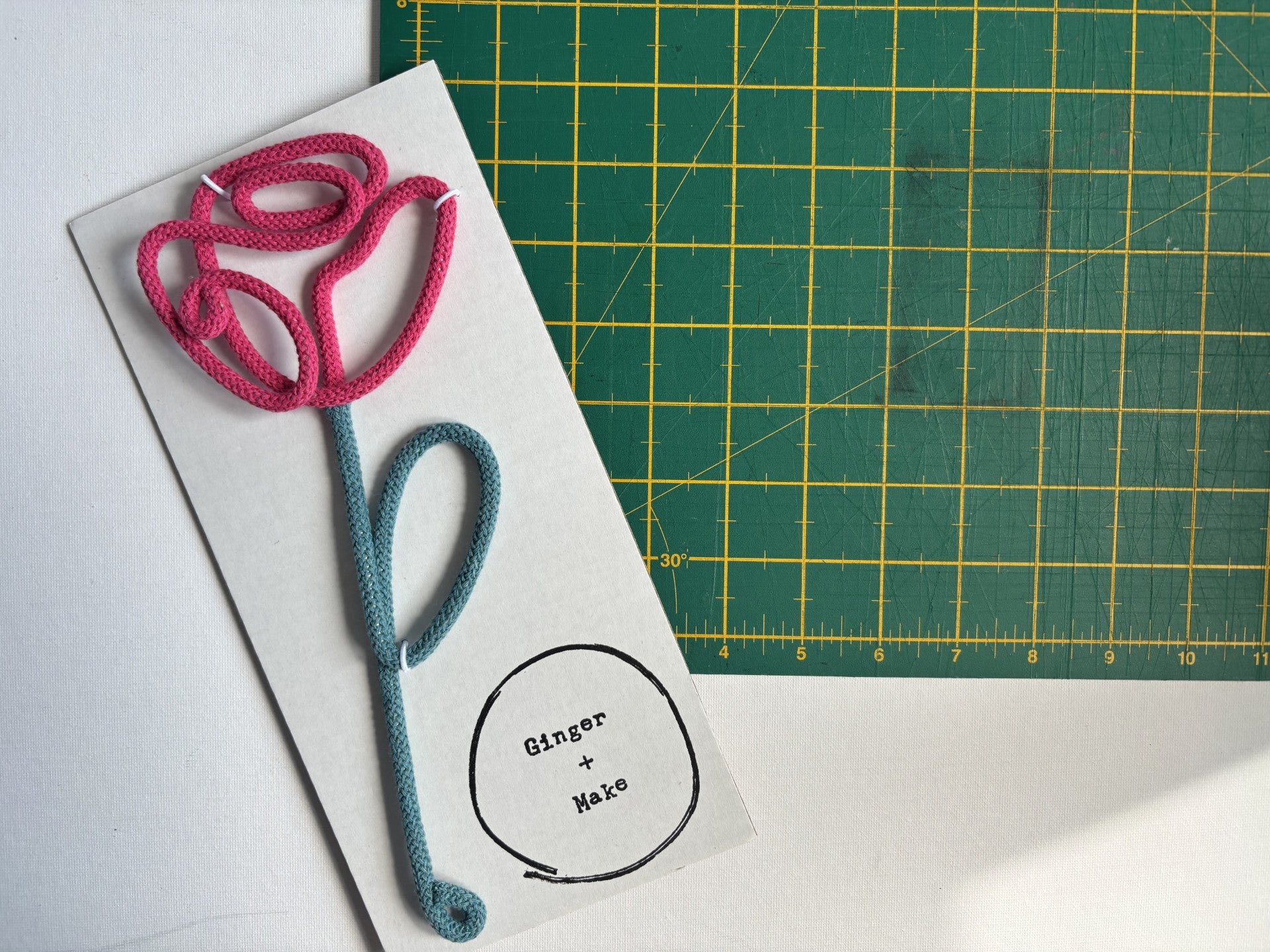 A handmade wire flower with cotton cord, with a pink top and teal stem on a white backing card that reads 'Ginger + Make'. A green cutting mat is visible in the background.