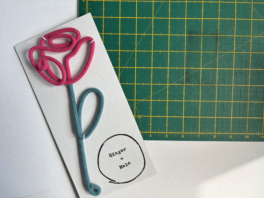A handmade wire flower with cotton cord, with a pink top and teal stem on a white backing card that reads 'Ginger + Make'. A green cutting mat is visible in the background.