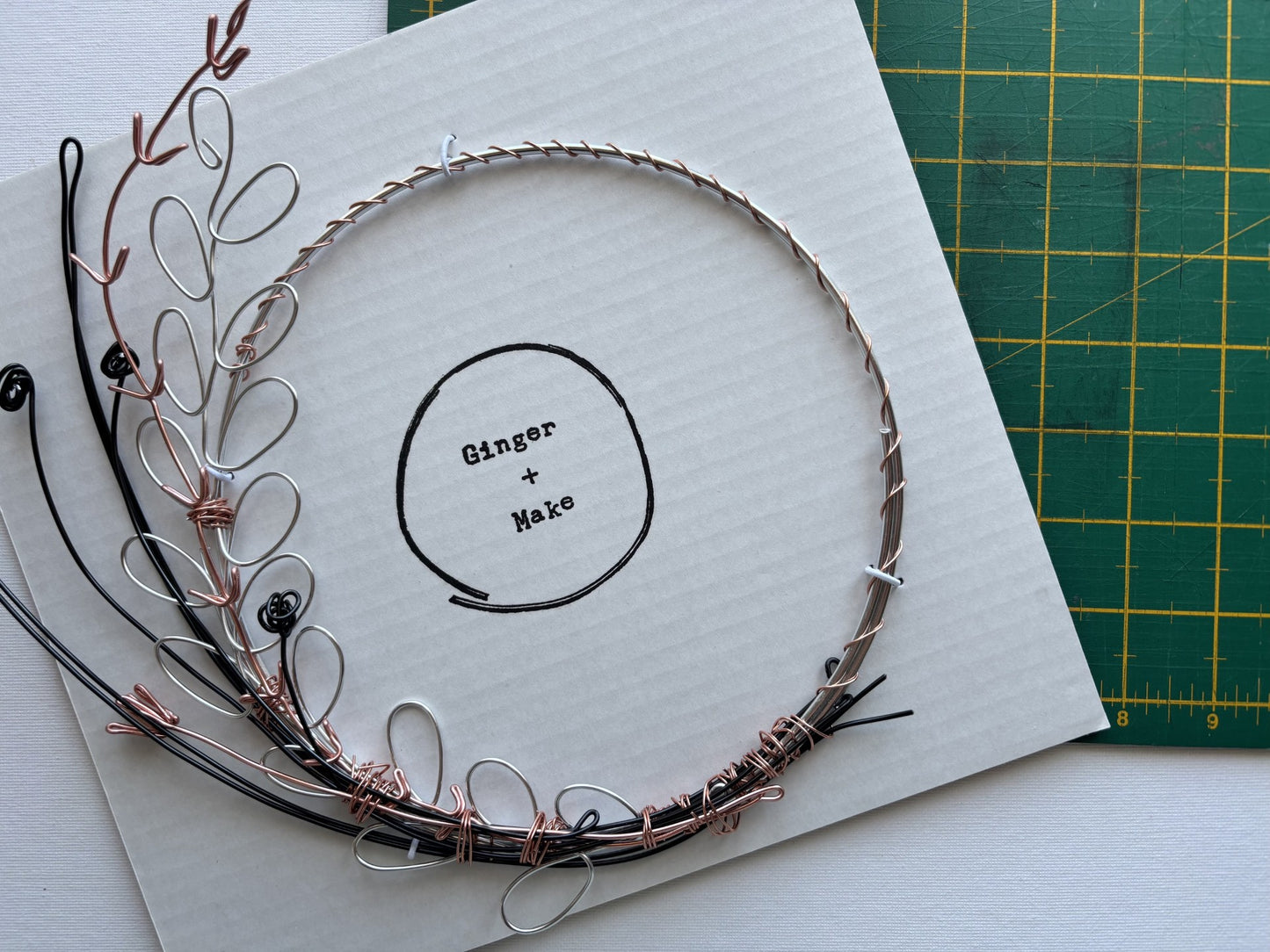 A handmade wire wreath with mixed metallic wire in silver, copper, and black, displayed on a white card that reads 'Ginger + Make'. A green cutting mat is visible in the background.