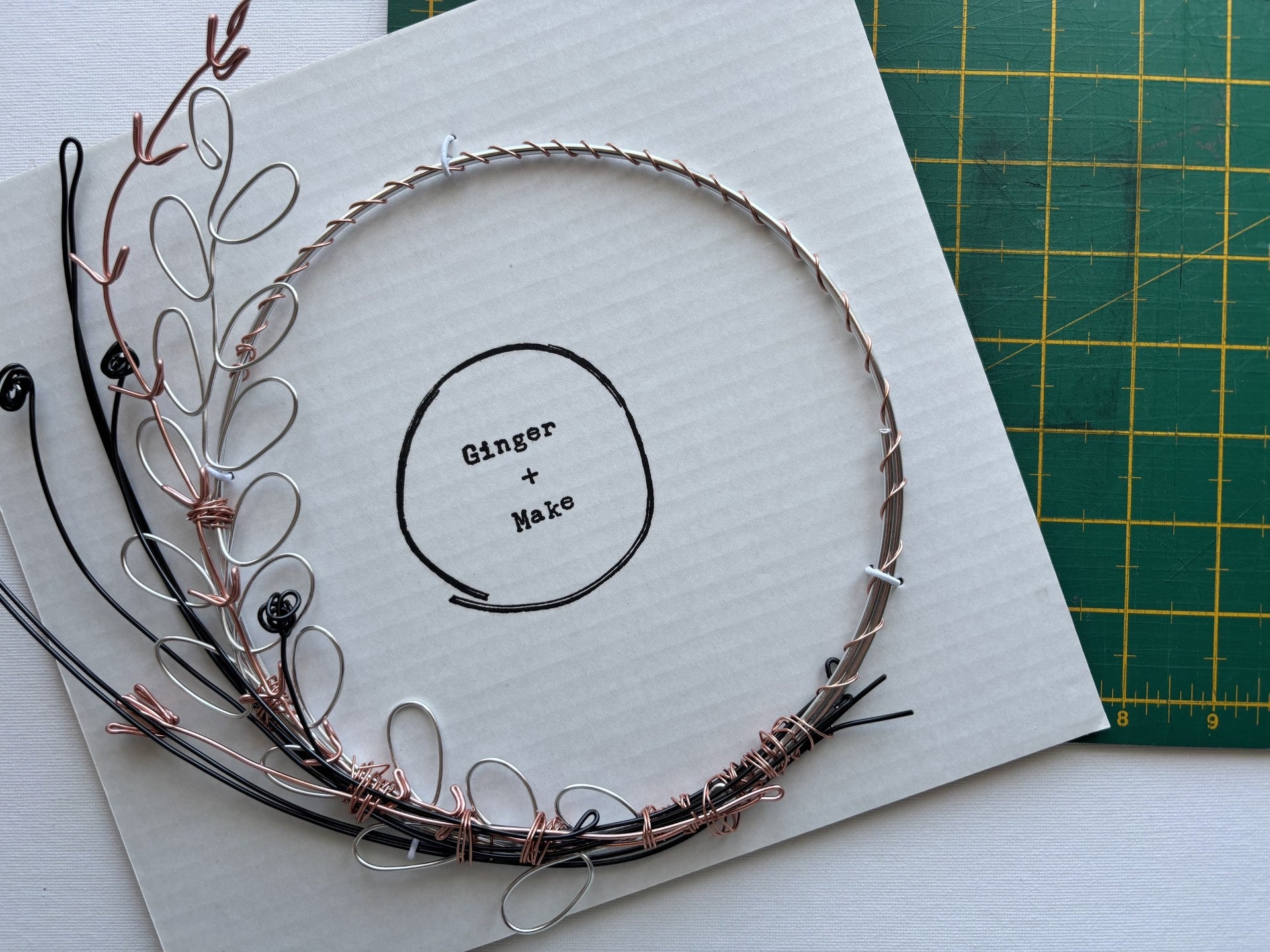 A handmade wire wreath with mixed metallic wire in silver, copper, and black, displayed on a white card that reads 'Ginger + Make'. A green cutting mat is visible in the background.