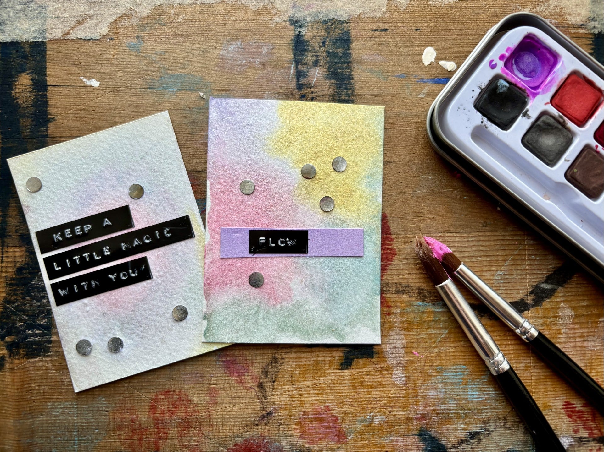 Two handmade watercolor cards displayed on a painted wooden surface, with a watercolor palette and paintbrushes nearby. One card is white with black text reading 'KEEP A LITTLE MAGIC WITH YOU!' and the other has a pastel watercolor wash with a purple band reading 'FLOW'.