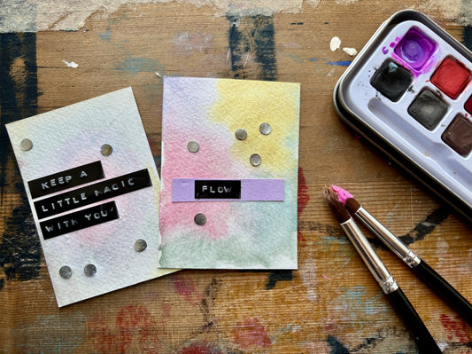 Two handmade watercolor cards displayed on a painted wooden surface, with a watercolor palette and paintbrushes nearby. One card is white with black text reading 'KEEP A LITTLE MAGIC WITH YOU!' and the other has a pastel watercolor wash with a purple band reading 'FLOW'.