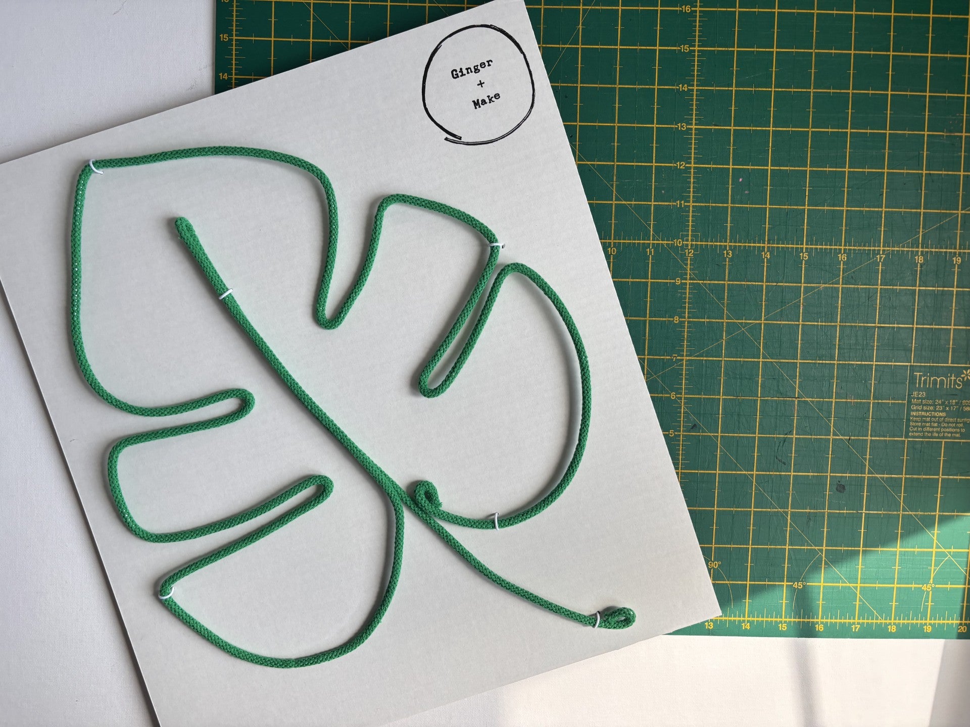 A green wire monstera leaf laid on a white square card, with a green cutting mat visible underneath.