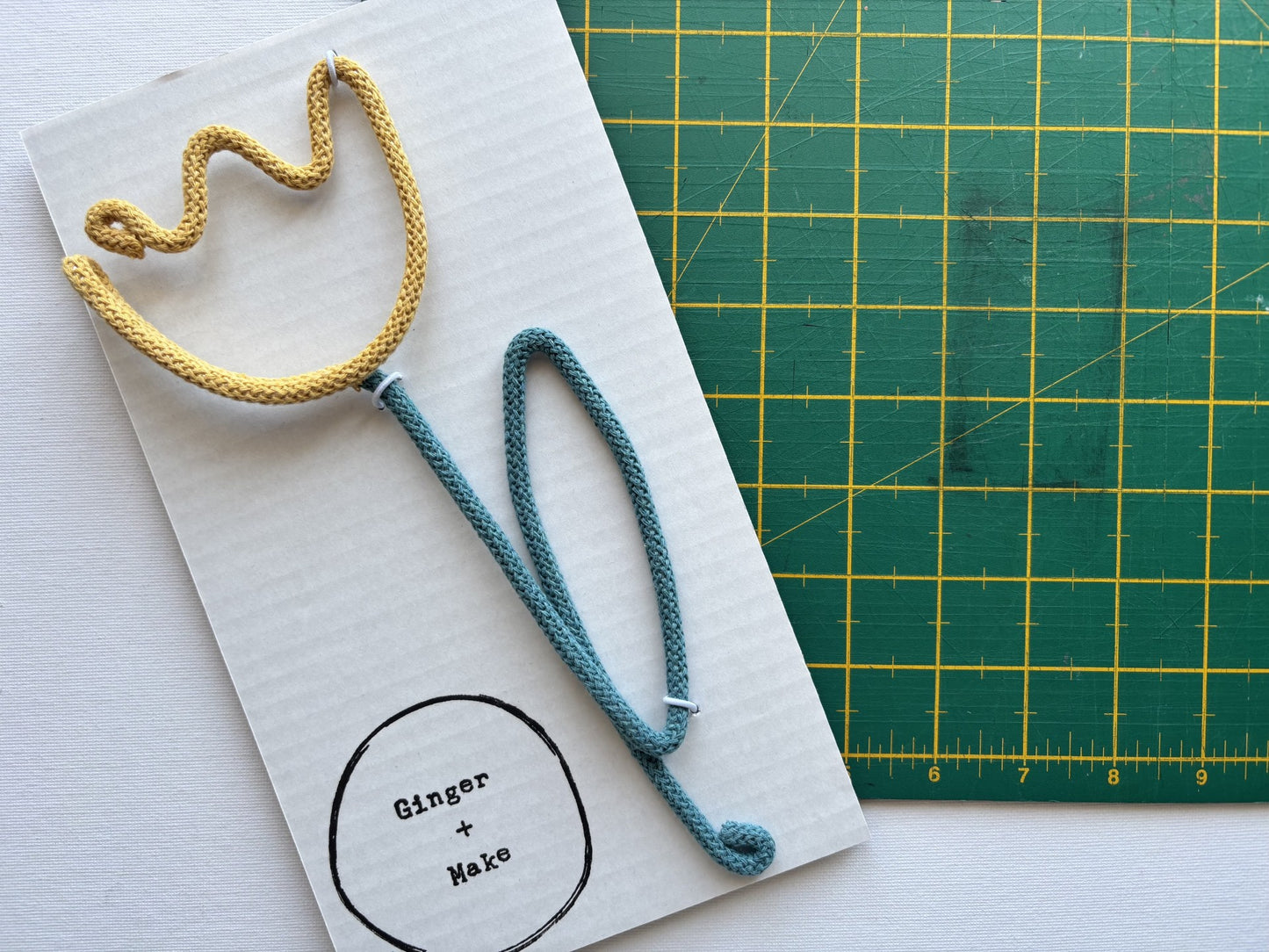 A handmade wire flower, with a yellow head and teal stem on white card, placed next to a green cutting mat.